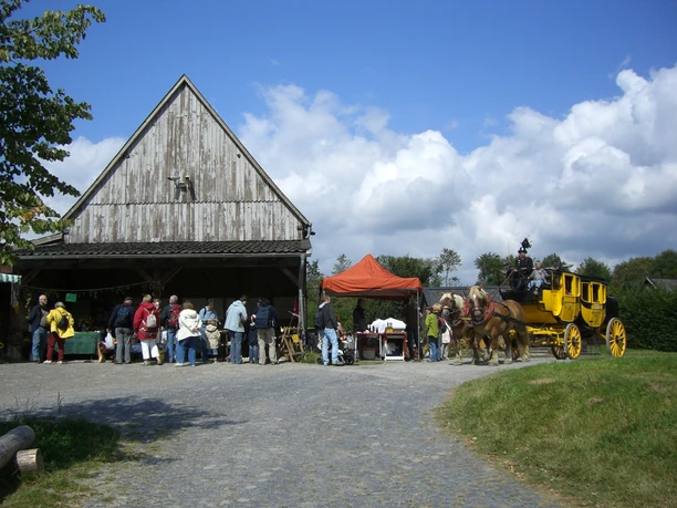 Bauernmarkt im Freilichtmuseum <p>Traditionelle Postkutsche vor historischem Fachwerkgebäude mit Besuchern auf steinigem Platz.</p>