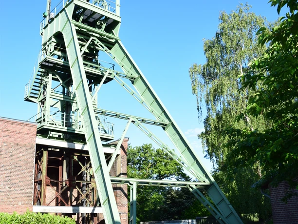 Förderturm der Grube Lüderich <p>Grüner Förderturm vor blauem Himmel, umgeben von Bäumen und Backsteingebäuden.</p>