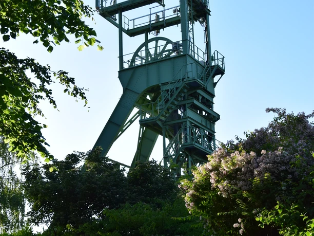 Förderturm der Grube Lüderich <p>Historisches Bergbaugerüst von Bäumen umrahmt, blauer Himmel im Hintergrund.</p>