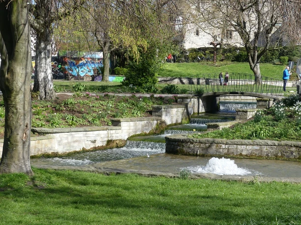 Brunnen fließt stufenweise über gemauerte Kaskaden, umgeben von blühenden Pflanzen und Gehwegen.