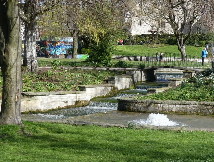 Brunnen fließt stufenweise über gemauerte Kaskaden, umgeben von blühenden Pflanzen und Gehwegen.