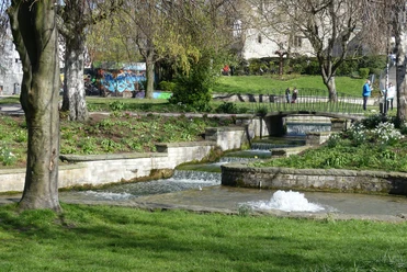 Paderquellgebiet - Kaskaden der Börnepader Brunnen fließt stufenweise über gemauerte Kaskaden, umgeben von blühenden Pflanzen und Gehwegen.