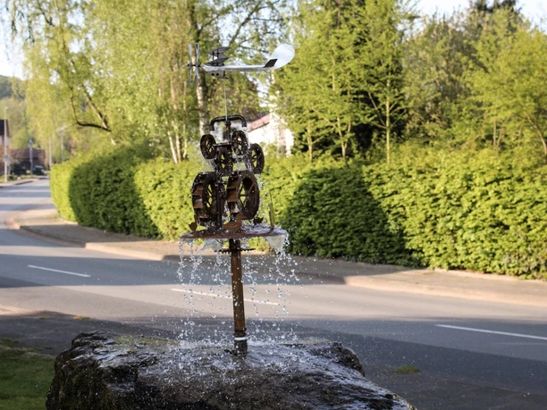 Wasserspiel Metallkonstruktion eines Wasserspiels auf einem Felsen am Straßenrand, umgeben von üppigem Grün.