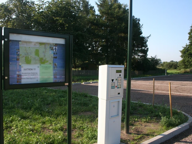Parkplatz mit Infotafel und Ticketautomat, im Hintergrund grüne Bäume unter klarem Himmel.