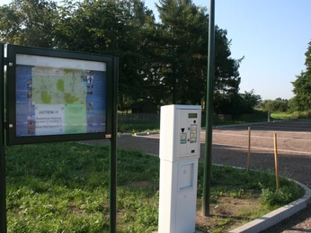 Wohnmobilstellplatz Rehburg Parkplatz mit Infotafel und Ticketautomat, im Hintergrund grüne Bäume unter klarem Himmel.