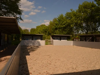 Großer Sandspielplatz mit überdachten Sitzbereichen, umgeben von Bäumen und klarem Himmel.