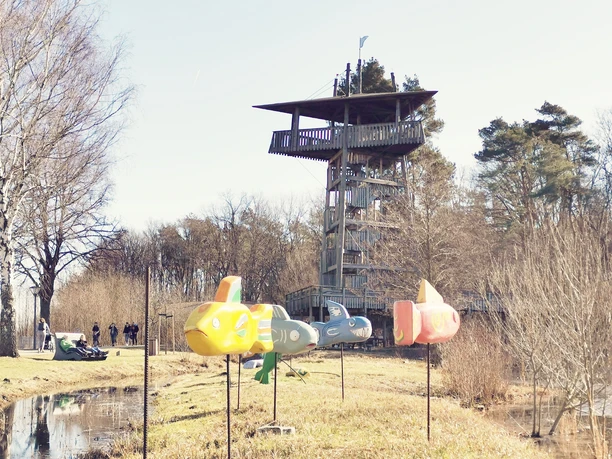 Holzbeobachtungsturm in Naturpark, umgeben von Bäumen, mit bunten Spielzeugflugzeugen im Vordergrund.
