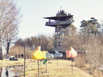 Blick auf den Aussichtsturm im Gartenschaupark Rietberg Holzbeobachtungsturm in Naturpark, umgeben von Bäumen, mit bunten Spielzeugflugzeugen im Vordergrund.