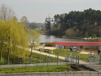 Blick vom Haupteingang auf den Aussichtsturm Ein See mit Strandkörben am Ufer, ein Beobachtungsturm und dichte Bäume im Hintergrund.
