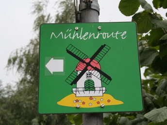 Schild Mühlentour Grünes Straßenschild mit Mühle, bezeichnet die Mühlenroute, zeigt Richtung mit weißem Pfeil.Green road sign with mill, designates the mill route, shows direction with white arrow.Grønt vejskilt med mølle, angiver mølleruten, viser retning med hvid pil.Groen verkeersbord met molen, geeft de molenroute aan, toont richting met witte pijl.