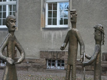 Familienskulptur Petershagen Drei bronzene Figuren, die in Interaktion stehen, vor einer Fassade mit Fenstern und einer Sockelmauer.