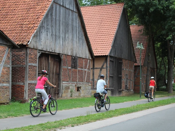 Drei Radfahrer passieren historische Fachwerkscheunen mit roten Ziegeldächern am Rand eines Weges.