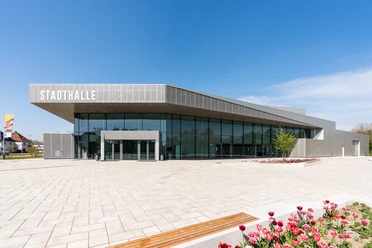 Moderne Stadthalle in elegantem Grau, umgeben von gepflastertem Platz und blühenden Tulpenbeeten.