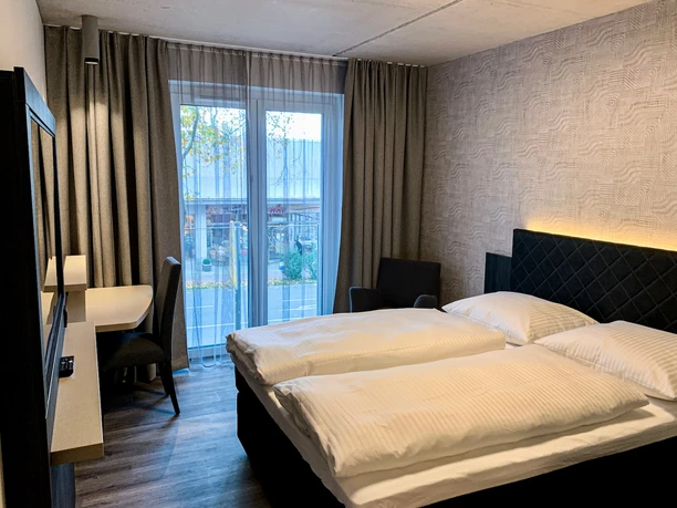 Hotel Zeitgeist - Doppelzimmer Doppelzimmer mit zwei Betten, Schreibtisch, Sessel und Fenster, das auf einen Außenbereich blickt.