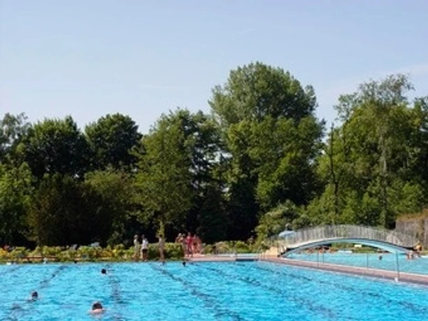Ein Freibad mit einem großen, blau schimmernden Becken, umgeben von üppigen Bäumen und einer Brücke.