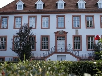 Historisches weißes Gebäude mit rotem Dach, umgeben von gepflegtem Grün und blühenden Rosen.