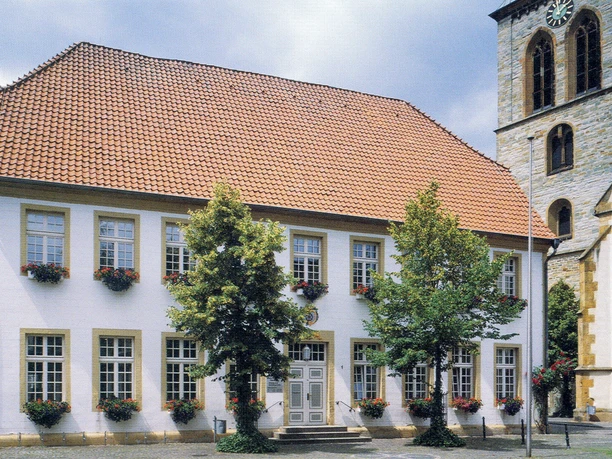 Historisches Gebäude mit rotem Ziegeldach, vielen Fenstern und Baumreihe, neben einer Kirche.