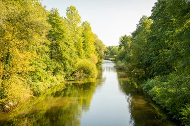 Aggerbogen Fluss fließt durch bewaldete Landschaft, Brücke im Hintergrund, sonniger Tag mit klarem Himmel.