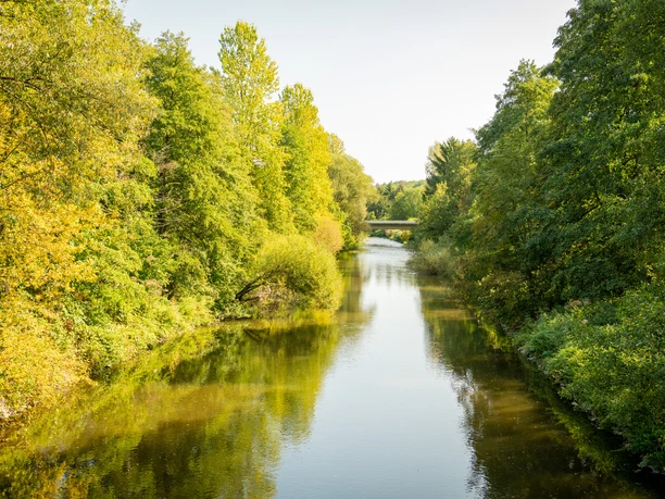 Aggerbogen Fluss fließt durch bewaldete Landschaft, Brücke im Hintergrund, sonniger Tag mit klarem Himmel.