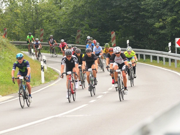 luedenbach-rennrad-mountainbike-tour-hotel_2.jpeg Radfahrer in bunten Trikots fahren in einer großen Gruppe eine kurvige, bewaldete Landstraße entlang.