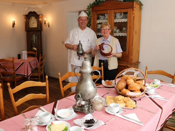 035 Krop. Berg. Kaffeetafel HN.JPG Ein älteres Paar in einer Gaststube hält einen Brotkorb, gedeckter Tisch mit Frühstück vor ihnen.
