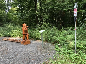 Märchenweg Bad Rehburg Holzskulptur eines Flötenspielers im Wald, umgeben von Schildern und üppigem Grün entlang eines Weges.