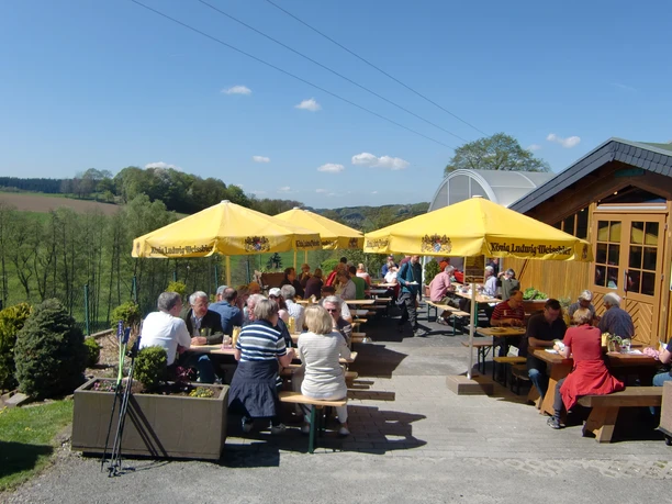 Biergarten-Tenne Menschen genießen bei sonnigem Wetter eine Mahlzeit auf einer Terrasse mit gelben Sonnenschirmen.