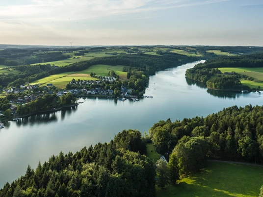 Bevertalsperre Luftbild einer natürlichen Flusslandschaft mit grünen Feldern, dichtem Wald und einer Siedlung am Ufer.