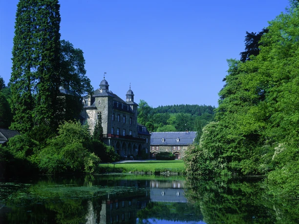 Schloss Gimborn Historisches Schloss mit zwei Türmen vor bewaldetem Hintergrund, spiegelt sich in einem ruhigen Teich.