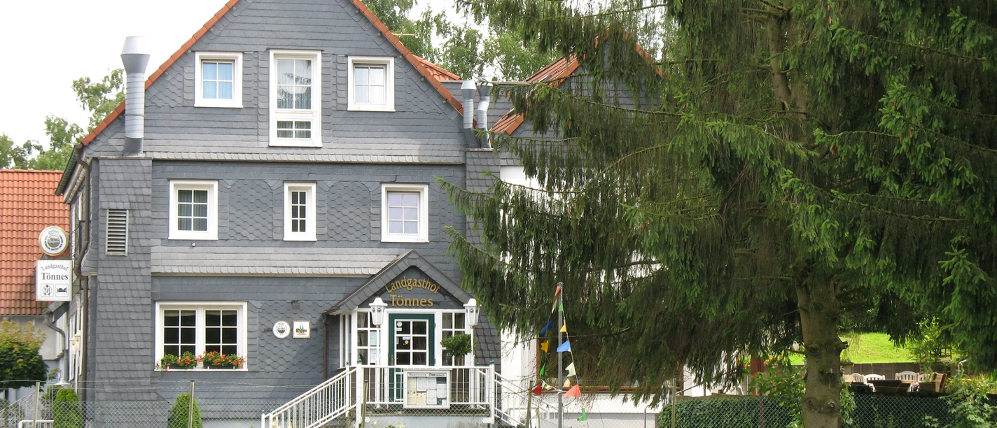 Landgasthof Tönnes Ein graues Fachwerkhaus mit roten Dachziegeln im Grünen, von Bäumen umgeben.