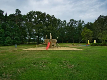 Spielplatz Offener Spielplatz auf einer Wiese, im Zentrum ein hölzernes Klettergerüst mit roter Rutsche, umgeben von Bäumen.