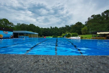 Schwimmbecken Ein glitzerndes, blaues Schwimmbecken im Freien, umgeben von Bäumen und Liegestühlen am Beckenrand.
