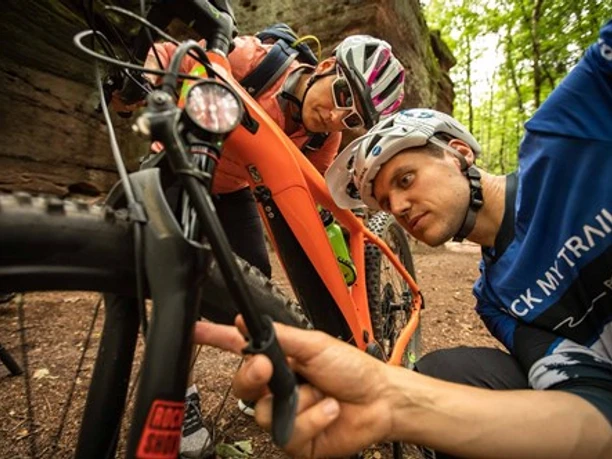 Rock My Trail Zwei Radfahrer inspizieren ihr Mountainbike im Wald, umgeben von Bäumen und Felsenformationen.