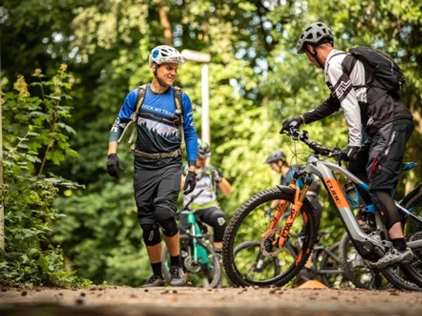 Rock My Trail Mountainbiker unterhalten sich neben ihren Fahrrädern im schattigen Wald in sportlicher Kleidung.