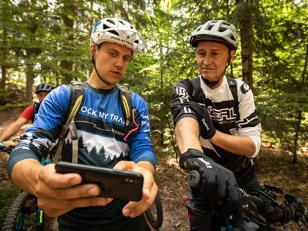 Rock My Trail Zwei Männer in sportlicher Kleidung schauen auf ein Smartphone, umgeben von einem Wald und Mountainbikes.