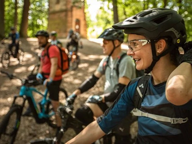 Rock My Trail Vier Radfahrer mit Helmen genießen eine Pause im schattigen Wald, dabei lachen und reden sie.
