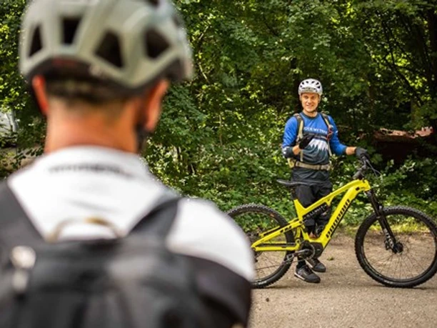 Rock My Trail Zwei Mountainbiker, einer im Vordergrund und einer lächelnd mit gelbem Fahrrad im Hintergrund, im Wald.