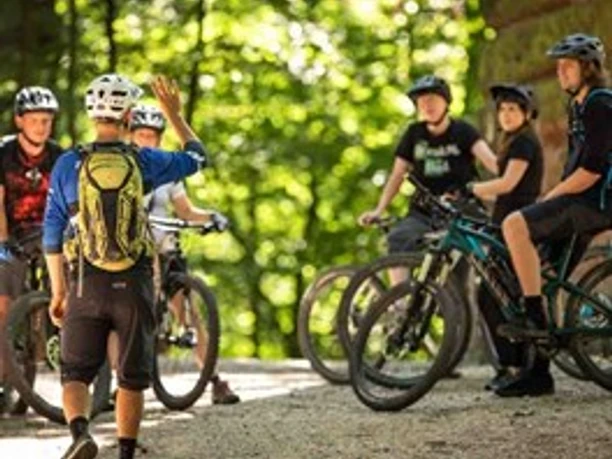 Rock My Trail Eine Gruppe von sechs Radfahrern mit Helmen und Rädern auf einem Waldweg, von Bäumen umgeben.