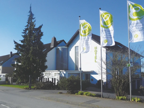 EH_Hotelansicht_c_EikamperHoehe.jpg Modernes Hotelgebäude mit blauen Akzenten, hohen Fenstern und wehenden Willkommensflaggen.