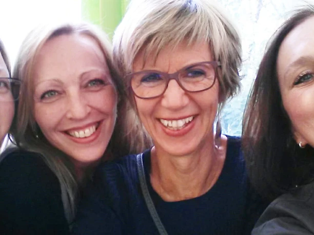 EH_Hotel_Team_c_EikamperHoehe.jpg Vier Frauen lächeln in die Kamera, eng beieinander, glücklich vor buntem Hintergrund.