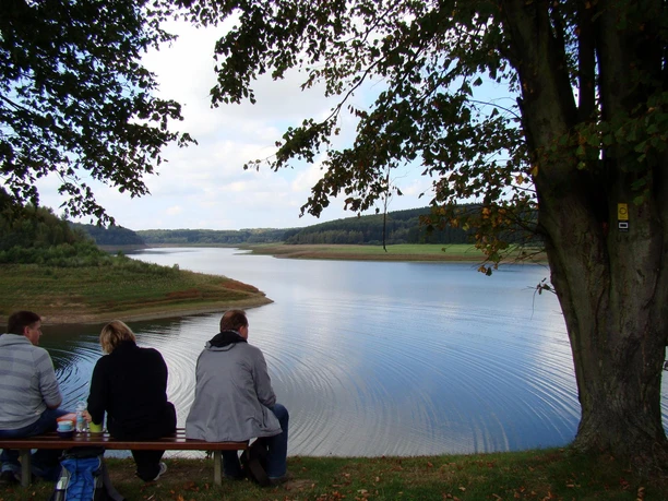 Panoramasteig-Et-3_MPU_140914_09.jfif Drei Personen sitzen auf einer Bank am Ufer eines ruhigen Sees, umgeben von grünen Hügeln.