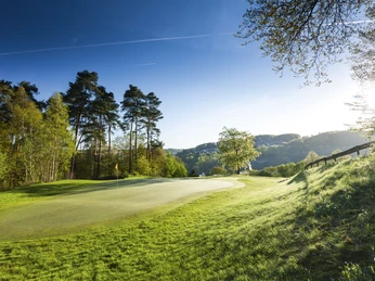 Golfclub Lüderich Golfplatz im Grünen mit Sonnenschein, Bäumen am Rand und Hügeln im Hintergrund.