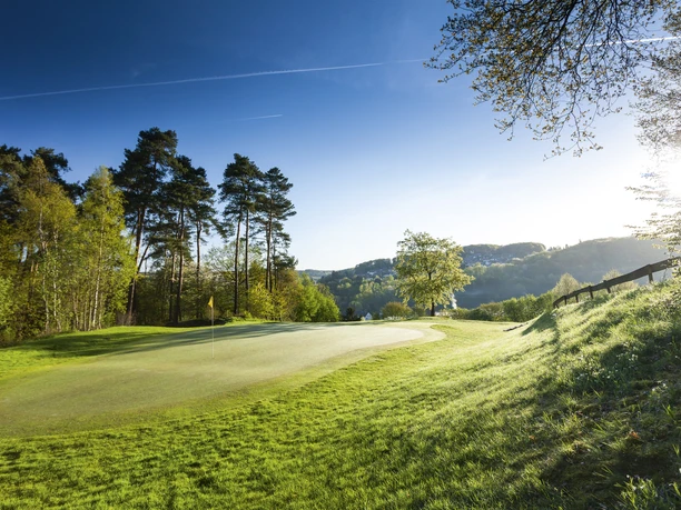 Golfclub Lüderich Golfplatz im Grünen mit Sonnenschein, Bäumen am Rand und Hügeln im Hintergrund.