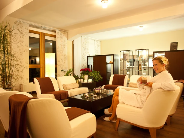 Beautyfarm.jpg Eine Frau im Bademantel entspannt mit Tee in einem stilvollen Spa-Wartebereich mit Sesseln.
