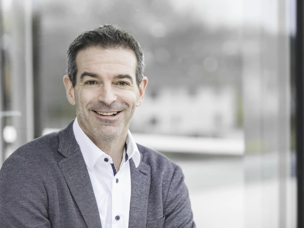 235_unbenannte-Fotosession_X3A0308.jpg Ein lächelnder Mann in Businesskleidung posiert vor verschwommenem Hintergrund, frohes Auftreten.