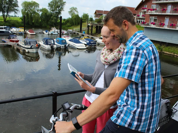 A couple on bicycles look at a map at Achim harbor with boats in the background.
