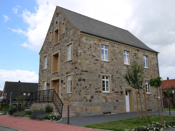 Museum Inselmühle Steingebäude mit Giebeldach und weißen Fenstern, umgeben von Blumenbeeten und neu gepflanzten Bäumen.