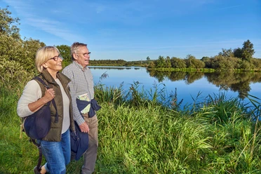 Rundwanderweg Heide Zwei Wandernde am Seeufer in grüner Landschaft