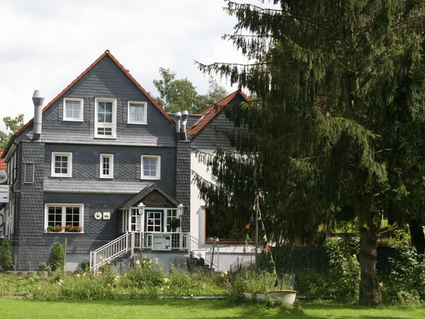 Aussenansicht_hinten.jfif Ein traditionelles zweistöckiges Gebäude mit Schieferfassade, umgeben von Bäumen und Wiesen.