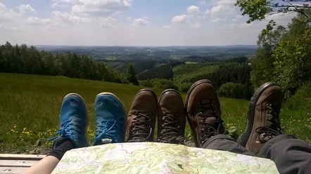 Reichshof-Blockhaus Drei Paar Schuhe und eine Landkarte auf einer Bank, mit weitem Blick auf bewaldete Hügel und Felder.